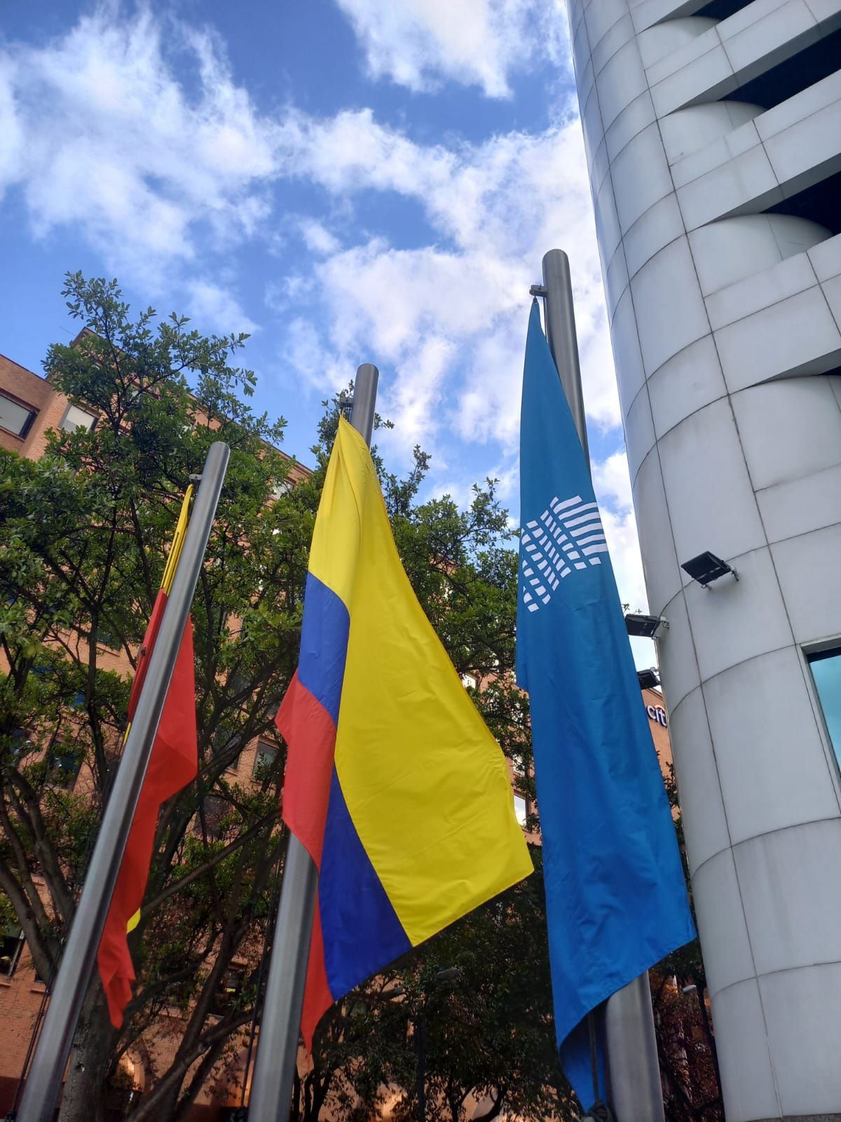 Three flags, including the Colombian tricolor and a blue UN flag, beside a white building and trees under a cloudy sky