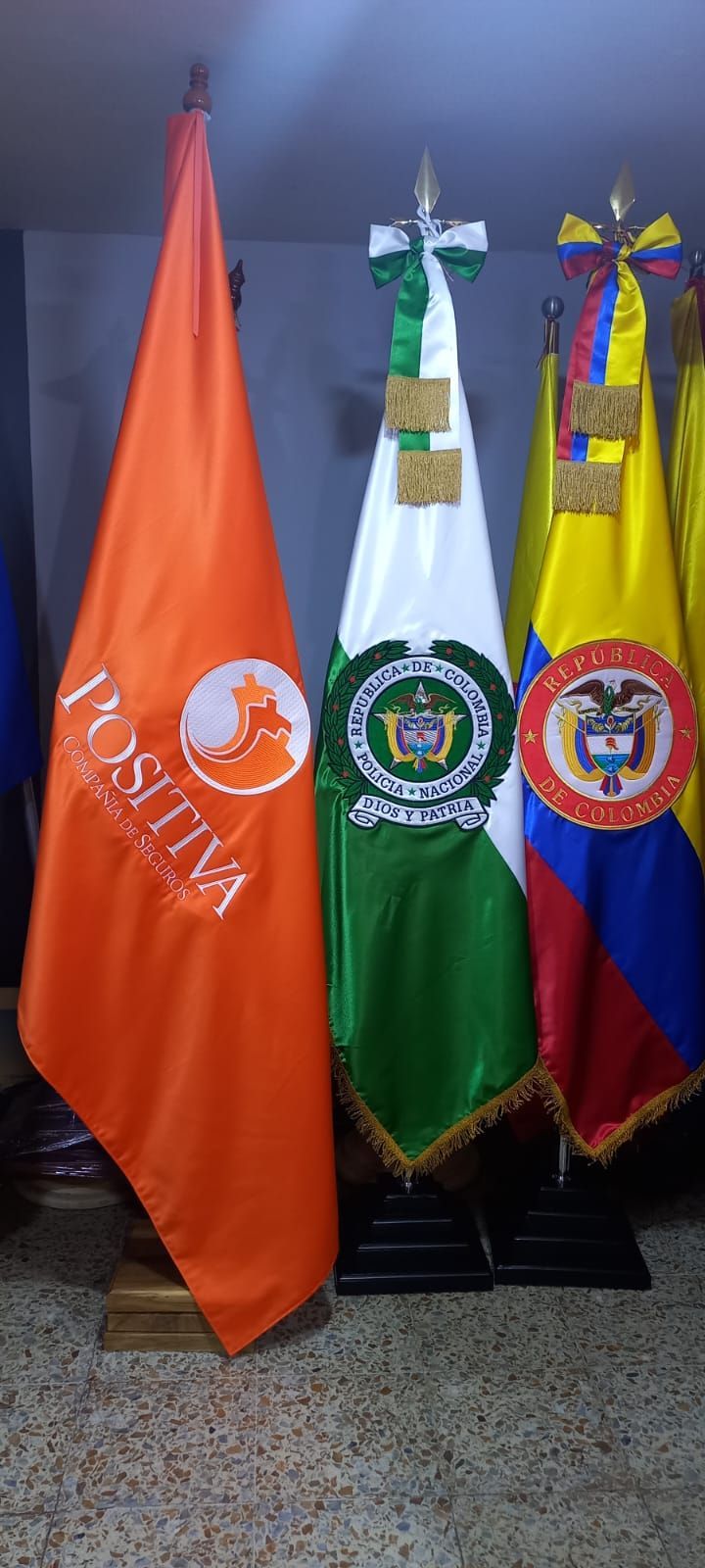 Three colorful institutional flags on stands against a wall, including orange, green, and blue-red-yellow flags.