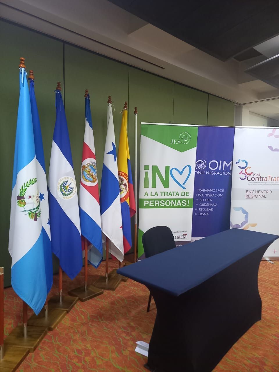 Escenario de la conferencia con banderas, una mesa cubierta con un mantel azul y pancartas de patrocinadores al fondo.