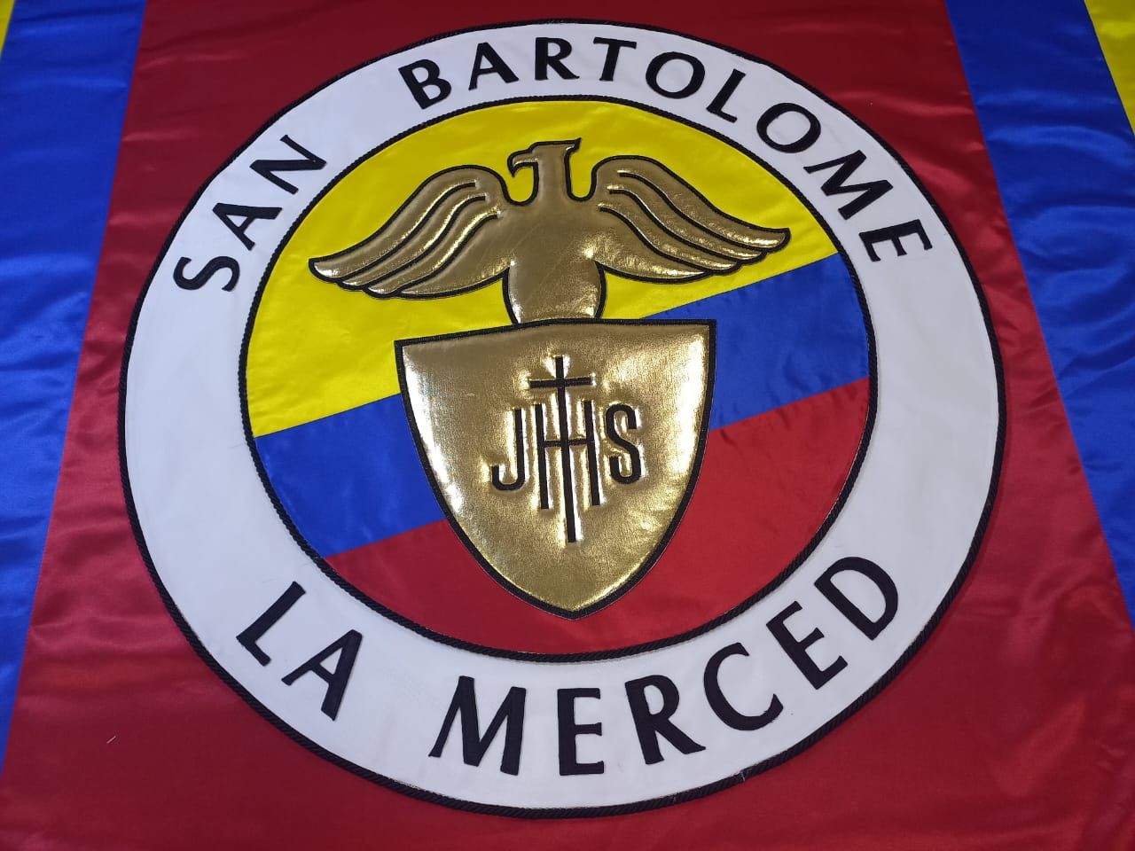 Emblema circular de San Bartolomé La Merced con águila y escudo dorados sobre fondo rojo, amarillo y azul.