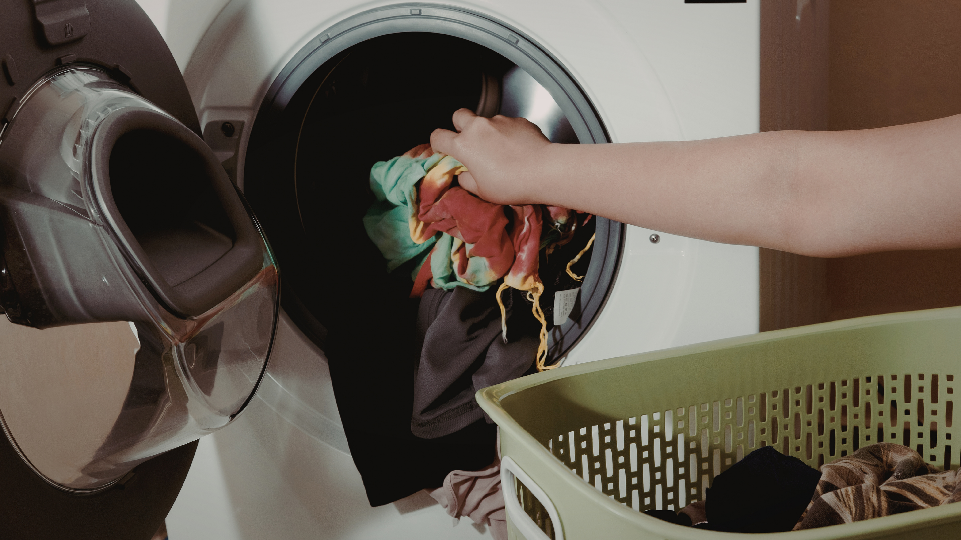 Person loading laundry into a washing machine, placing clothes next to a shoe and a laundry basket.