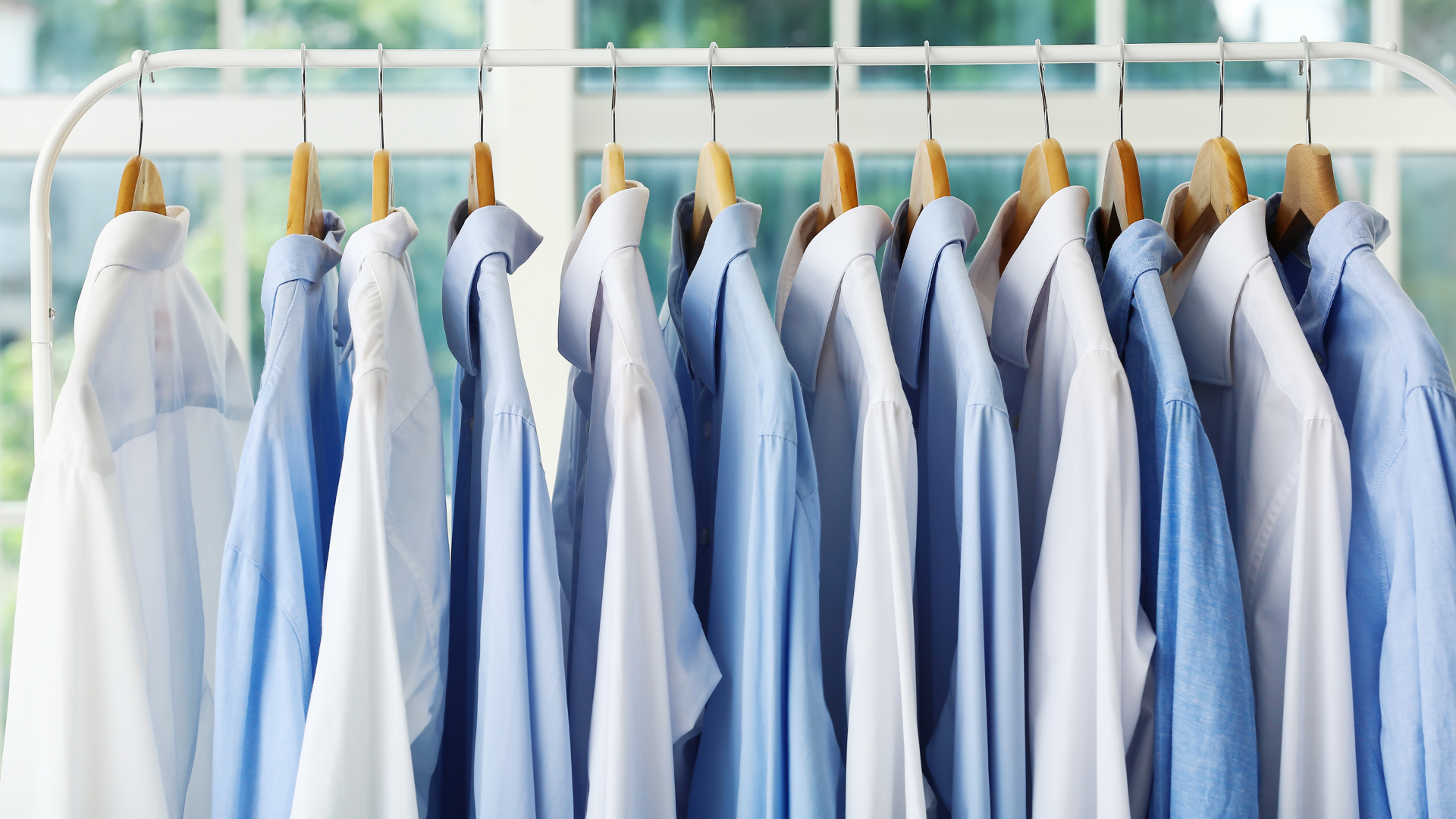 Blue and white button-up shirts hang on wooden hangers on a rack, near a window.