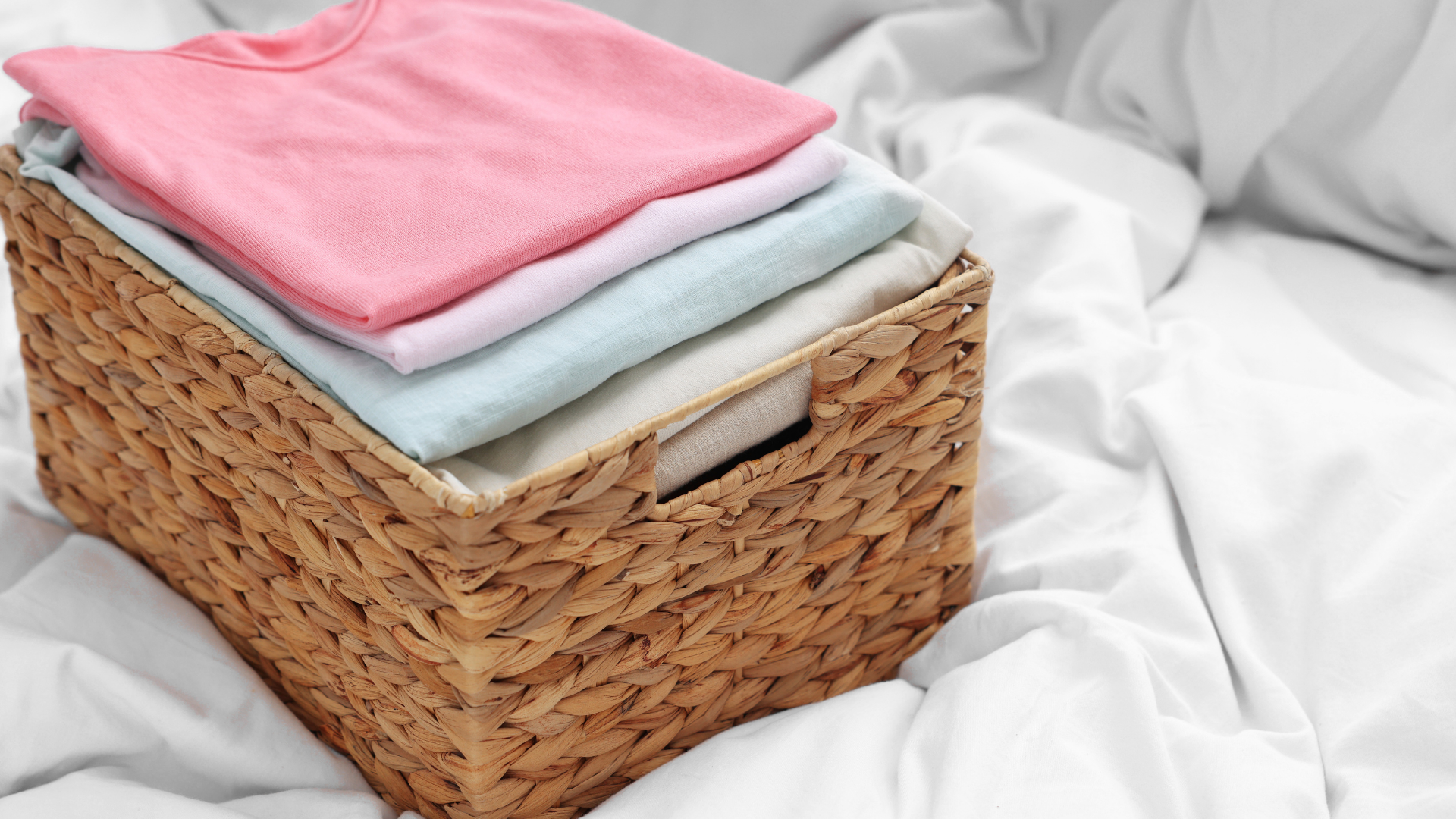 Wicker basket with folded pastel clothing on a white bed.