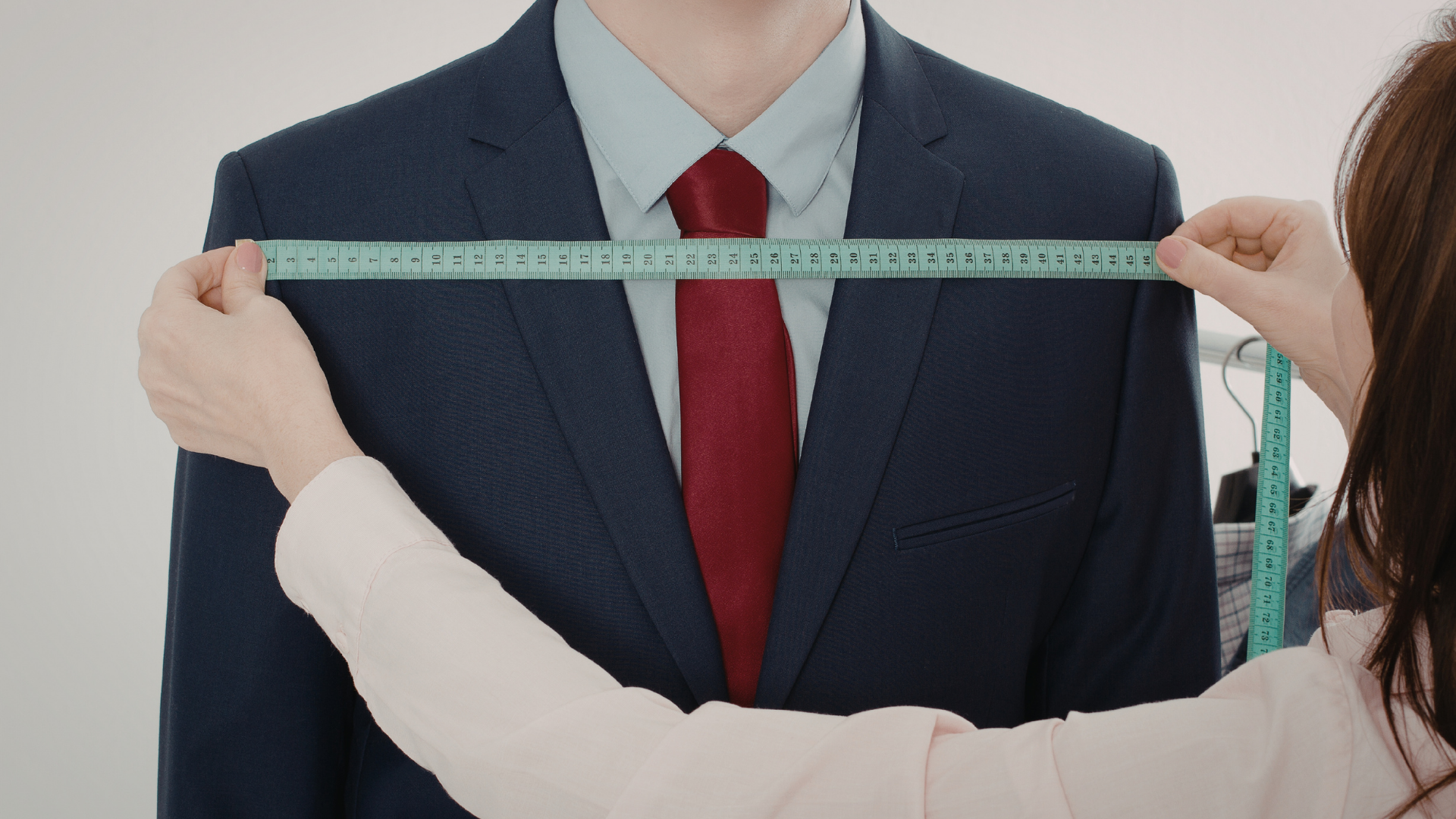 A person in a blue suit is measured across the shoulders by another person with a measuring tape.