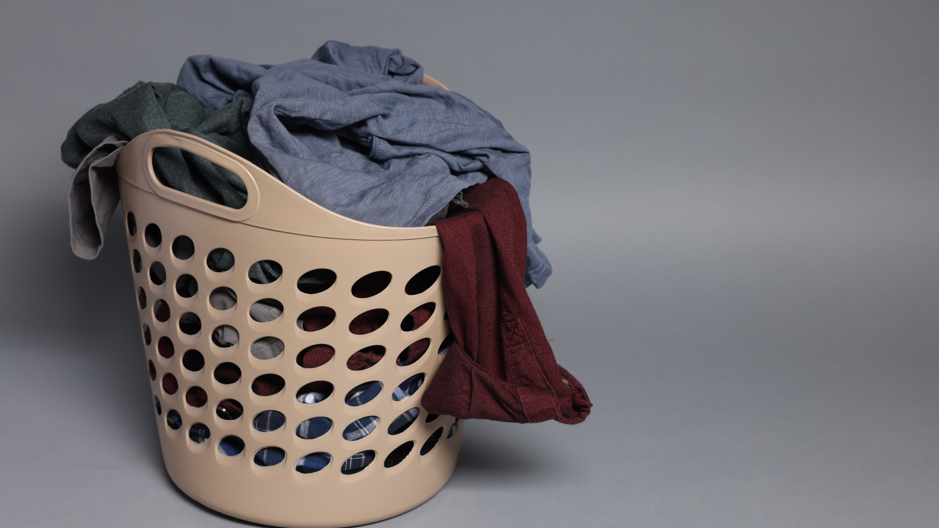 Laundry basket overflowing with blue, red, and gray clothing on a gray background.