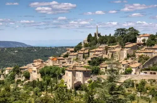 Village perché sur une colline, avec des maisons en pierre et un clocher dominant une vallée verdoyante sous un ciel bleu.