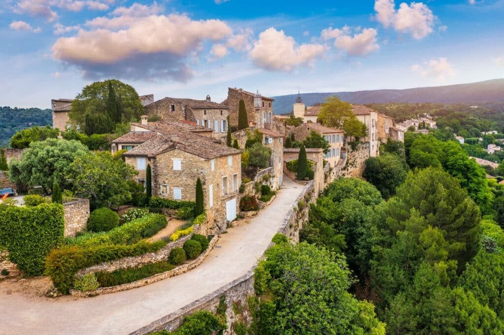 Village de pierre aux toits de tuiles rouges, le long d'une route sinueuse, entouré d'une végétation luxuriante sous un ciel bleu.