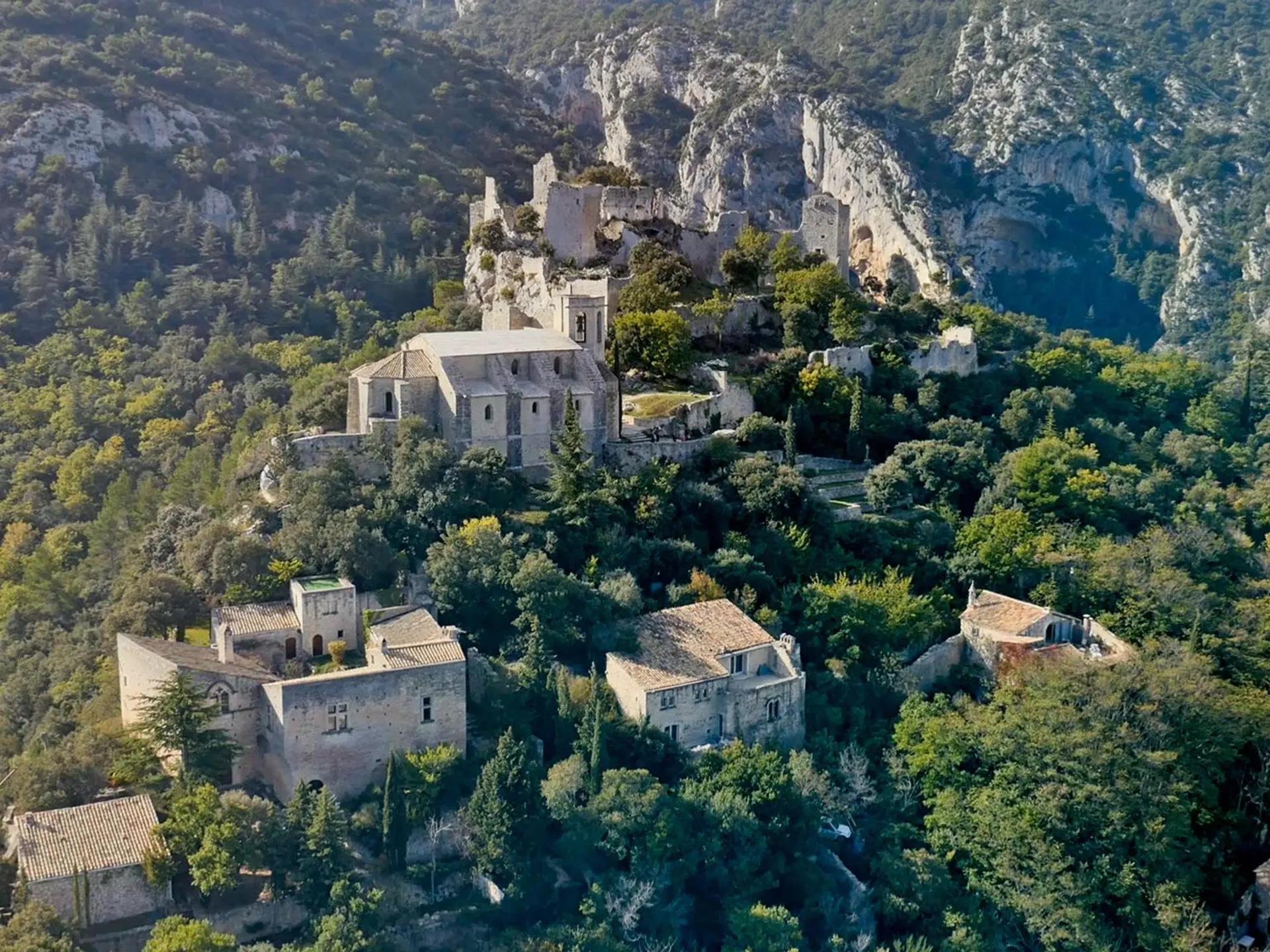 Village de pierre perché sur une colline, entouré d'une dense végétation verte, au pied de falaises blanches escarpées.
