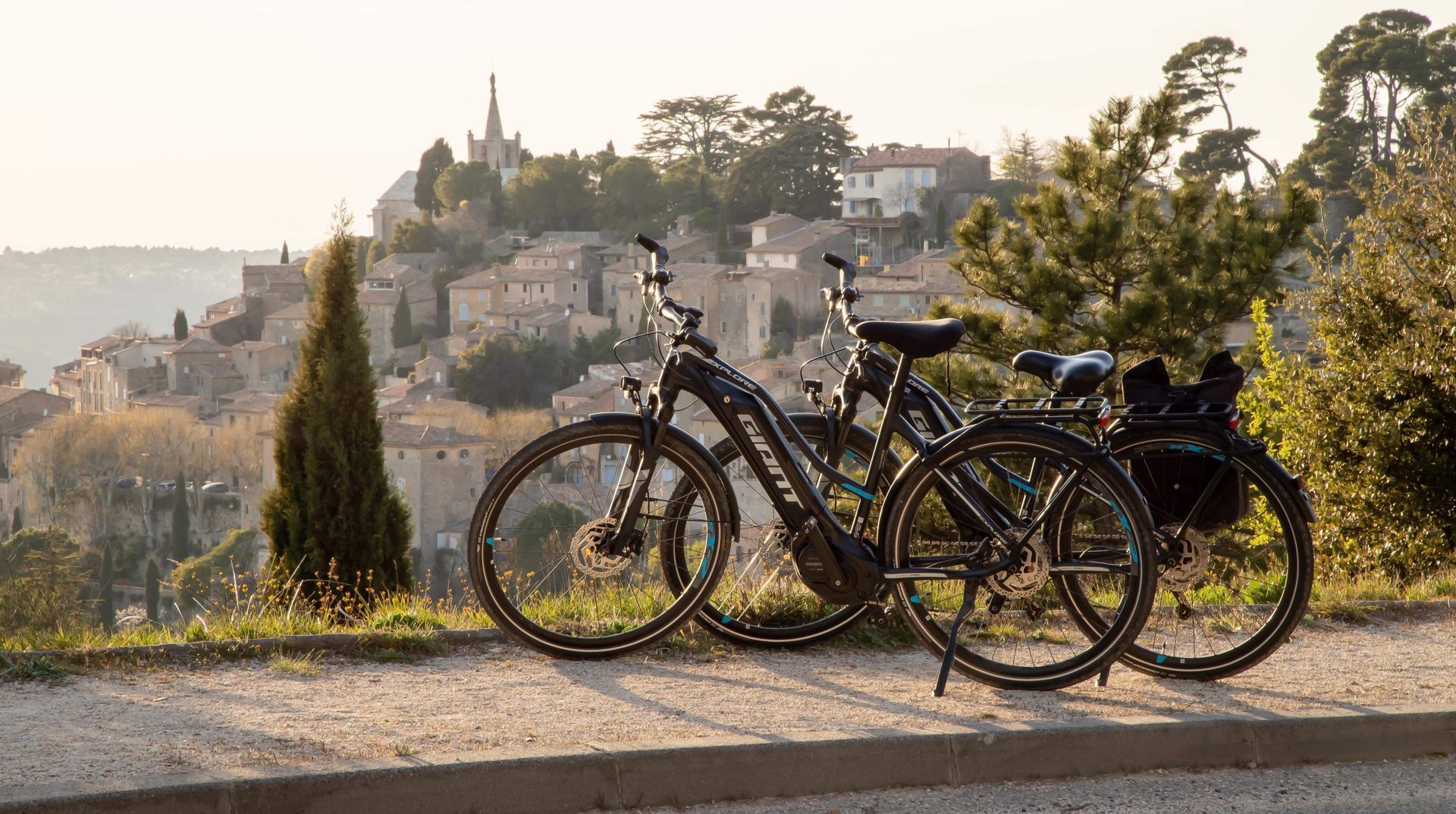 Deux vélos garés sur un promontoire au sommet d'une colline, avec des arbres et des bâtiments en arrière-plan, au coucher du soleil.