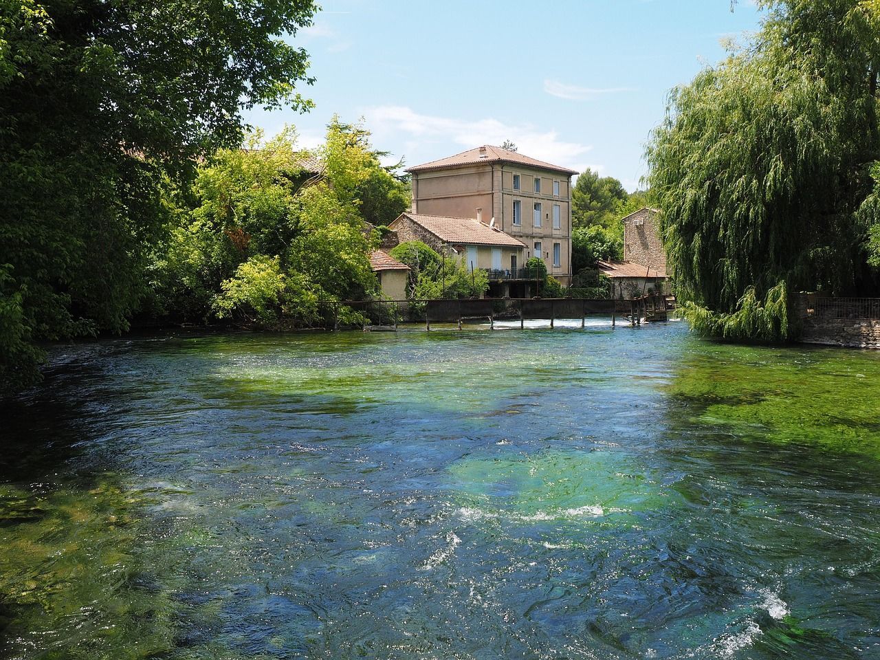Rivière aux eaux claires bordée d'arbres, avec un bâtiment en pierre en arrière-plan sous un ciel bleu.