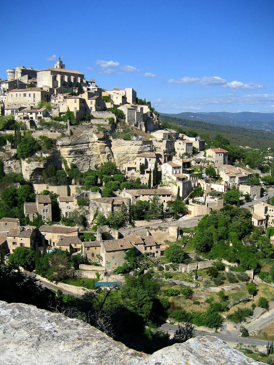 Village de pierre perché sur une colline, avec des maisons beiges regroupées, des terrasses verdoyantes et un ciel bleu en arrière-plan.