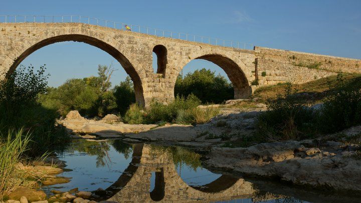 Pont en pierre arquée enjambant une rivière calme, où se reflètent les arbres et le ciel bleu dans l'eau.