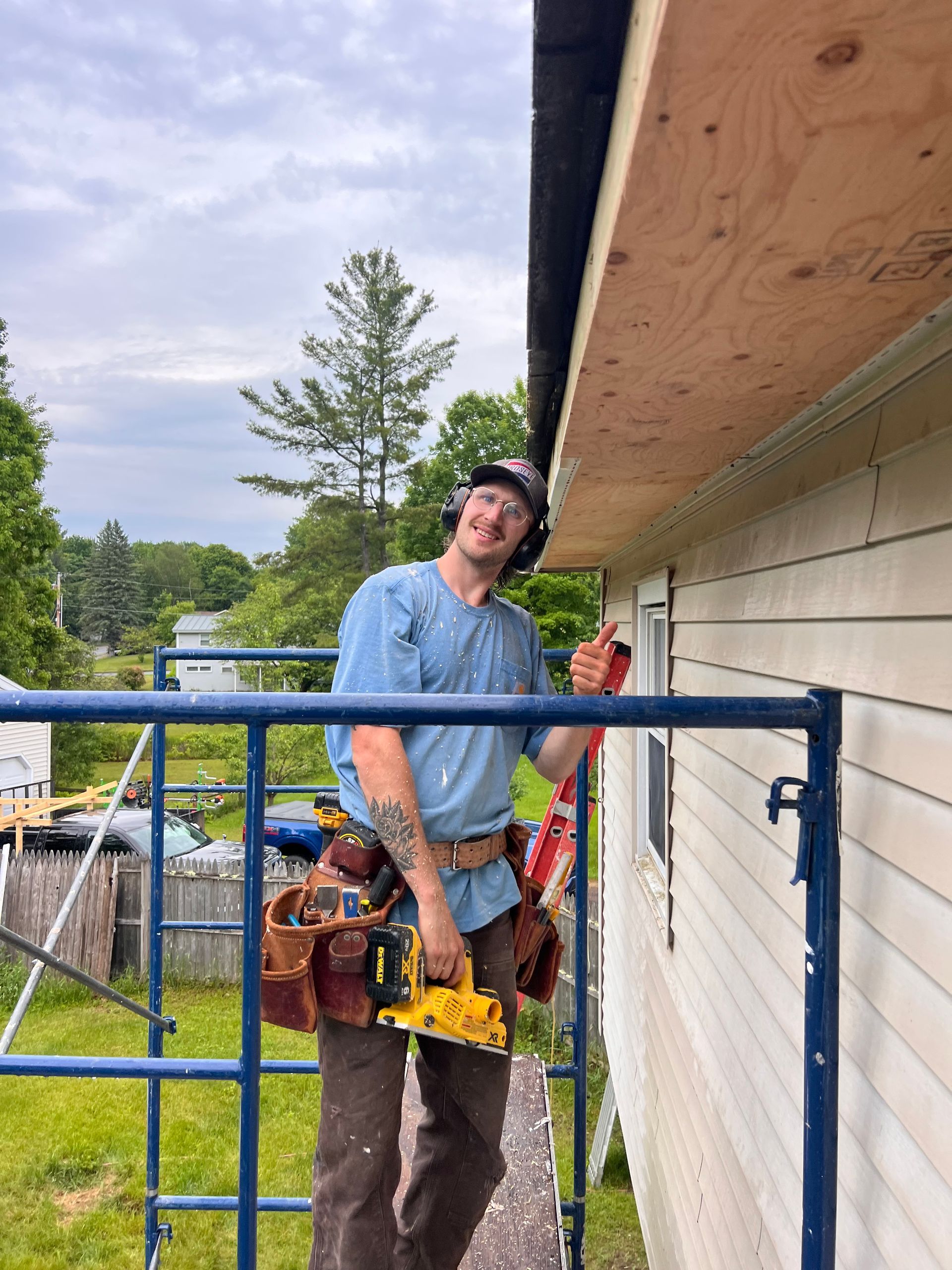 Worker in blue shirt using a yellow power tool on a house exterior by a blue railing.