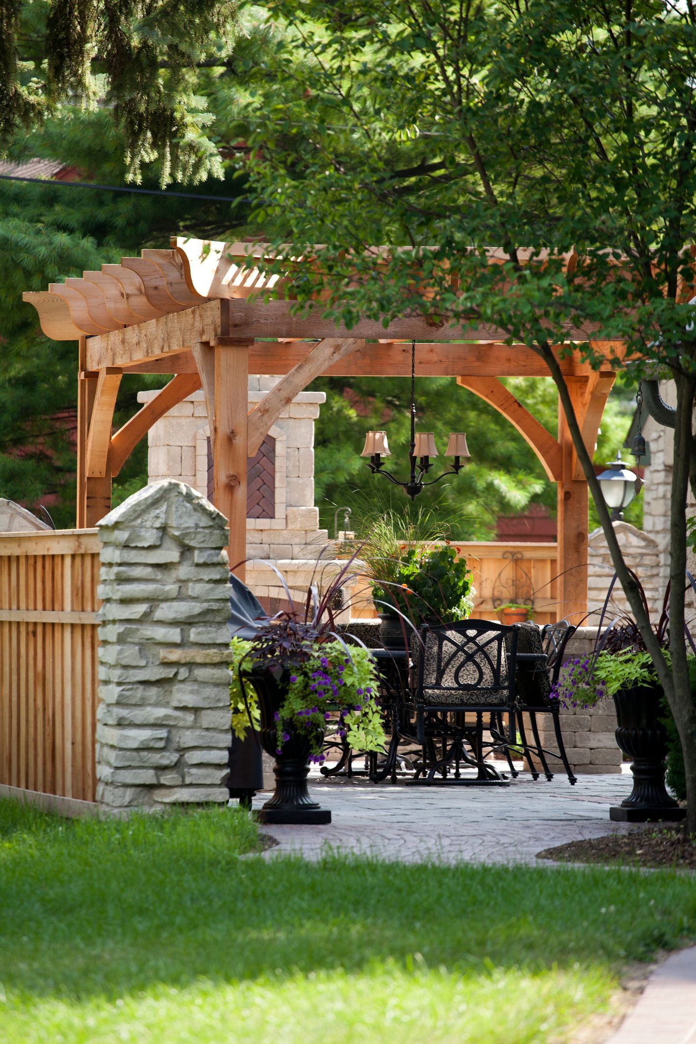 Backyard patio with wooden pergola, stone pillar, and black wrought-iron table set under leafy trees