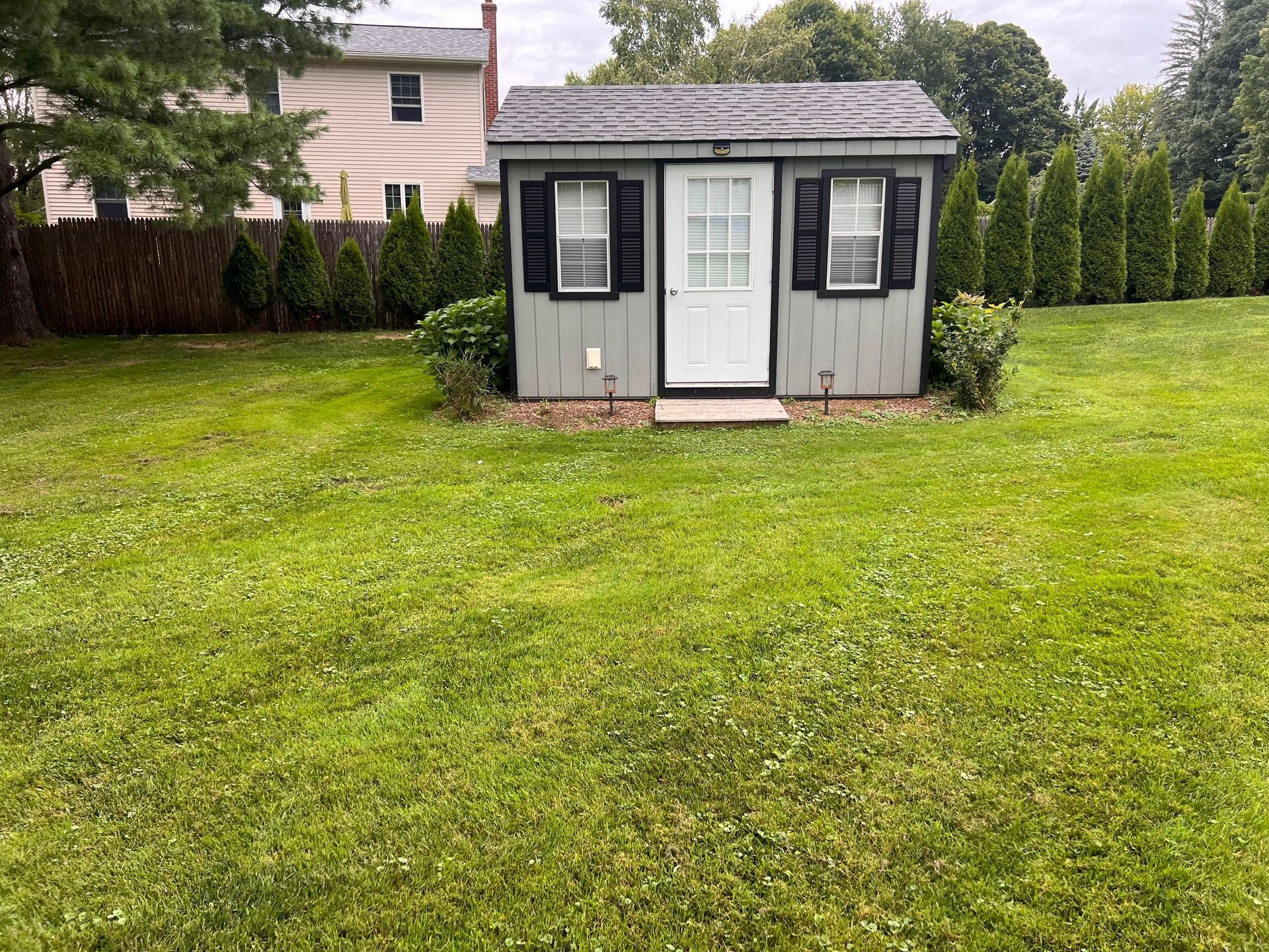 Small gray backyard shed with black shutters and white door on a grassy lawn.