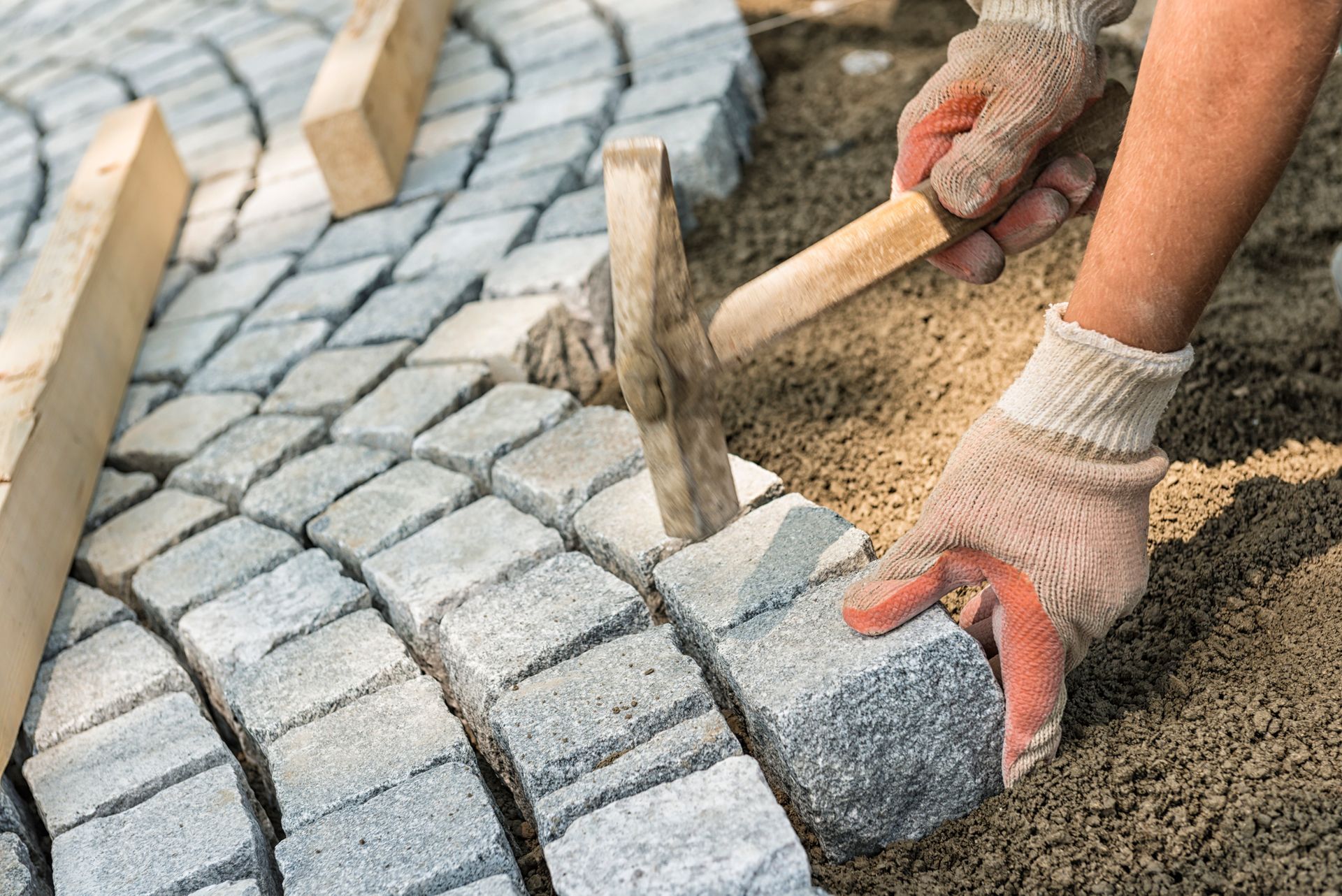Hands laying gray cobblestone pavers into sand during sidewalk installation