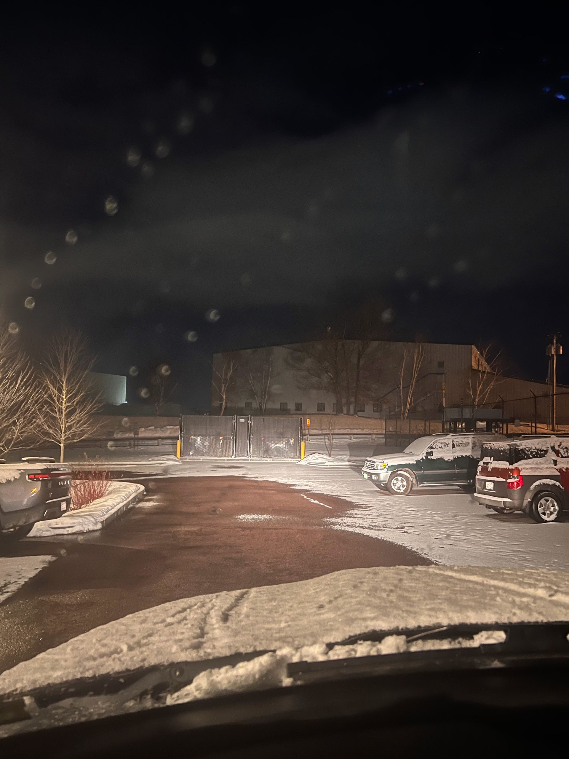Snowy parking lot at night with parked cars and a building in the background