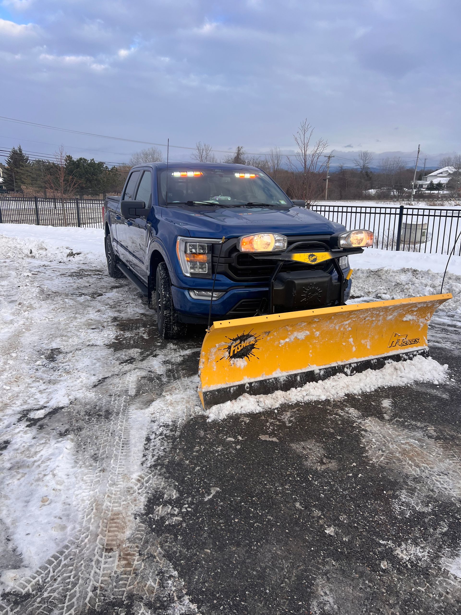 Blue pickup truck with a yellow snow plow parked on a snowy driveway