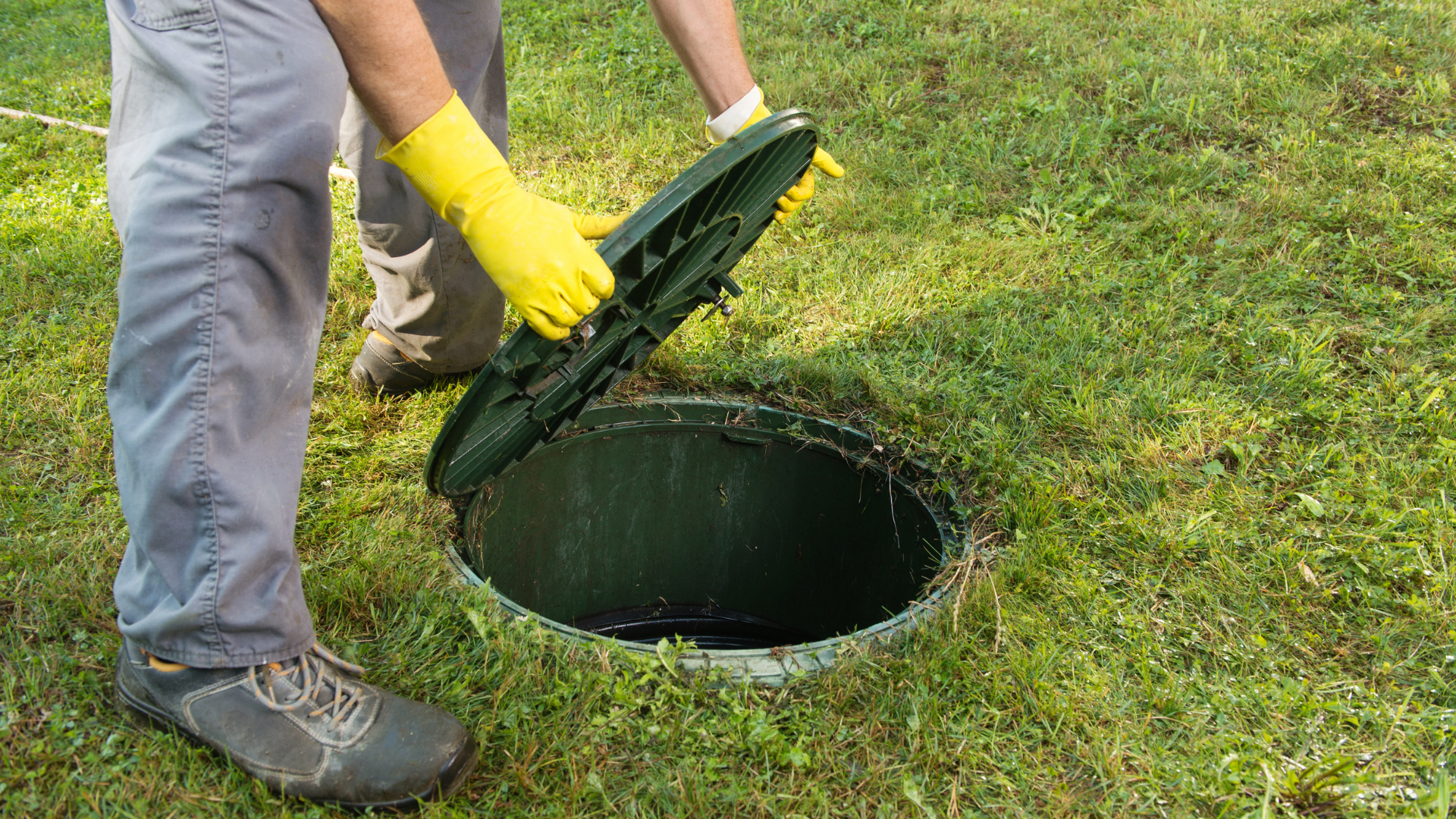 Person in yellow gloves lifting a manhole cover on a grassy lawn.