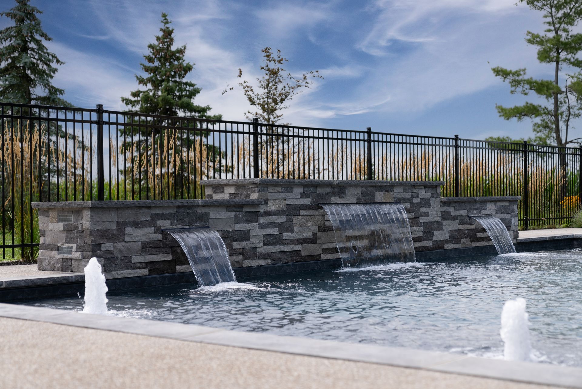 Tiered stone fountain with waterfalls into a pool, backed by a black fence and trees under a blue sky