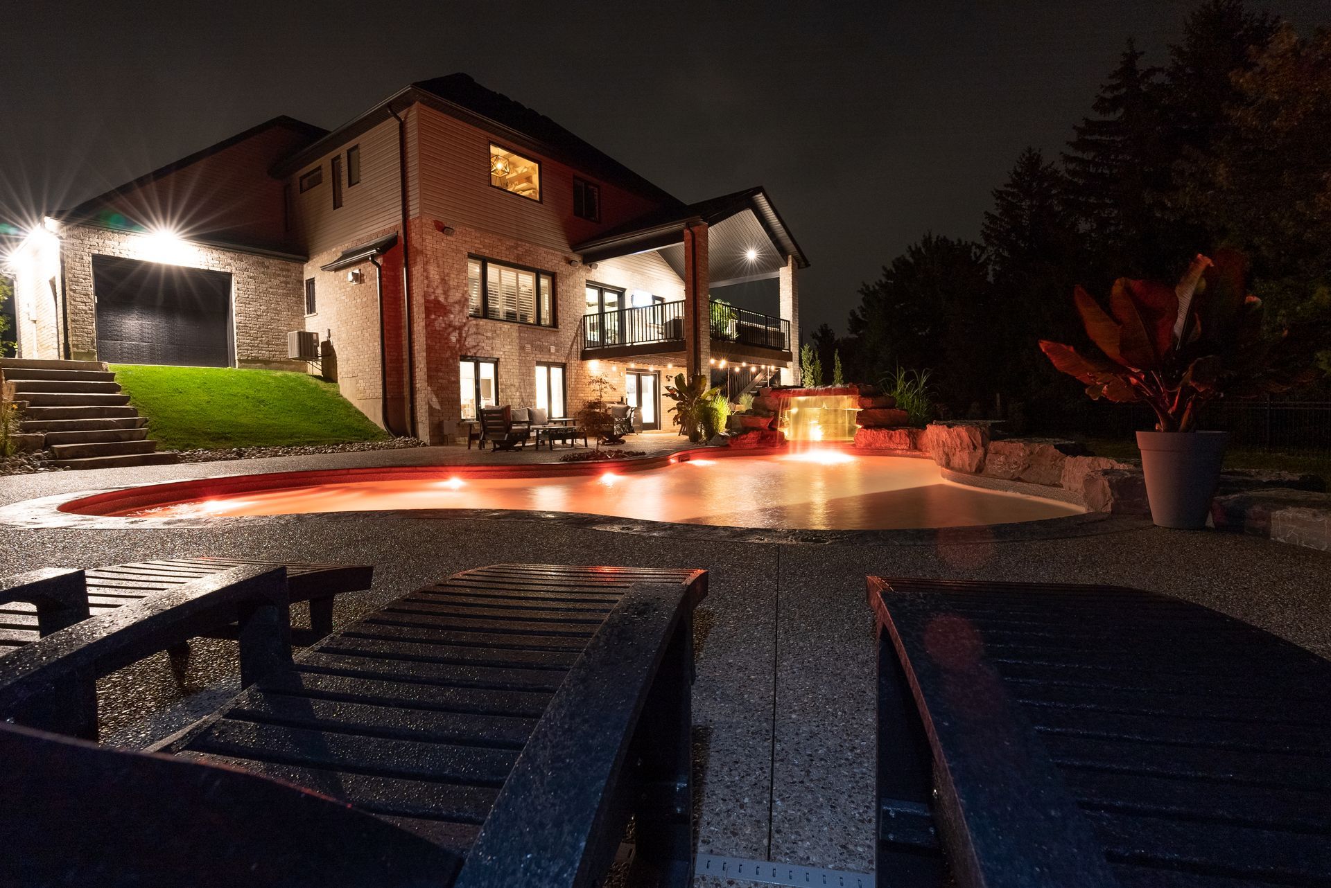 Nighttime view of a lit luxury house and pool with glowing red water and lounge chairs in the foreground