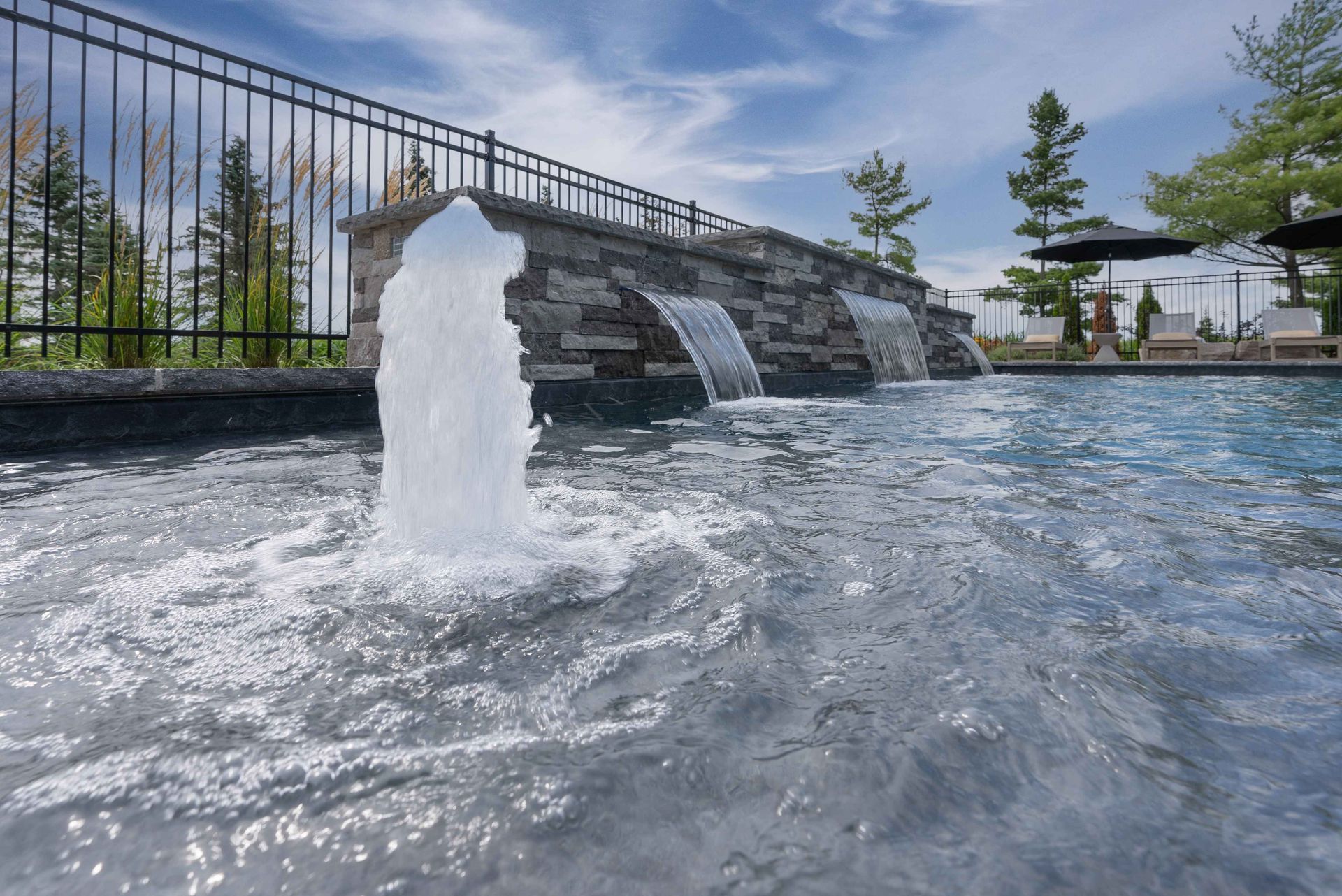 Modern pool with a water fountain, waterfall wall, and landscaped patio under a blue sky