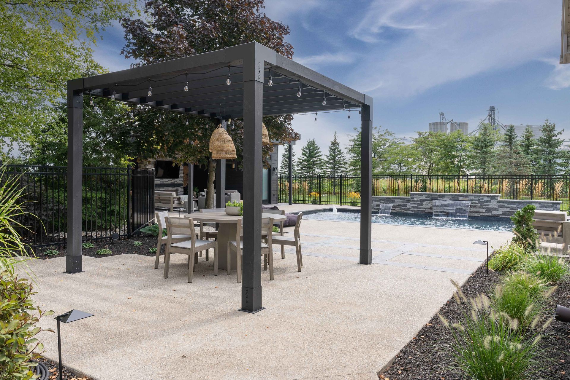 Outdoor patio with black pergola, dining table, and chairs beside a pool and landscaped garden.
