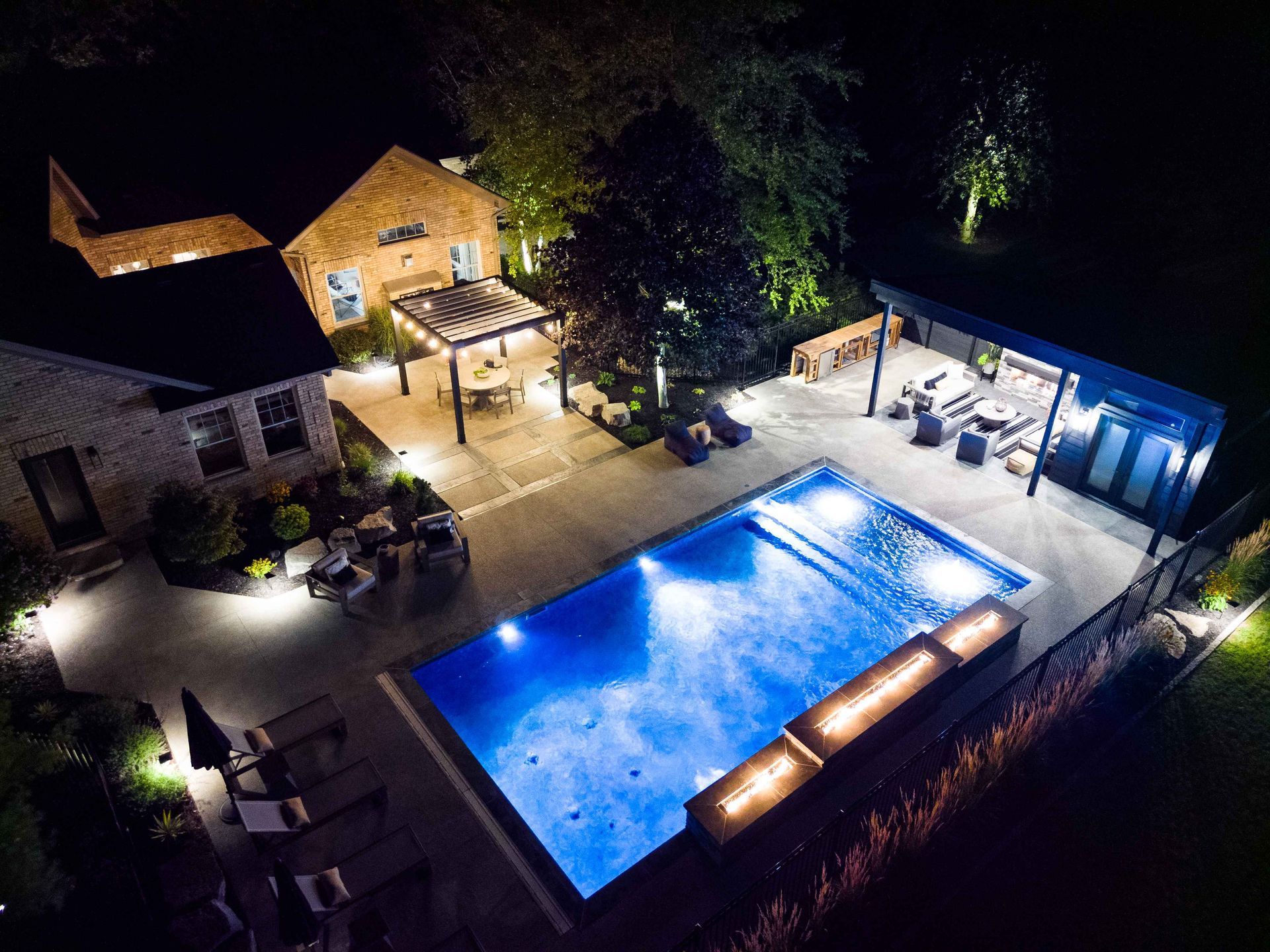 Nighttime aerial view of a lit backyard with a blue swimming pool, patio seating, and surrounding buildings
