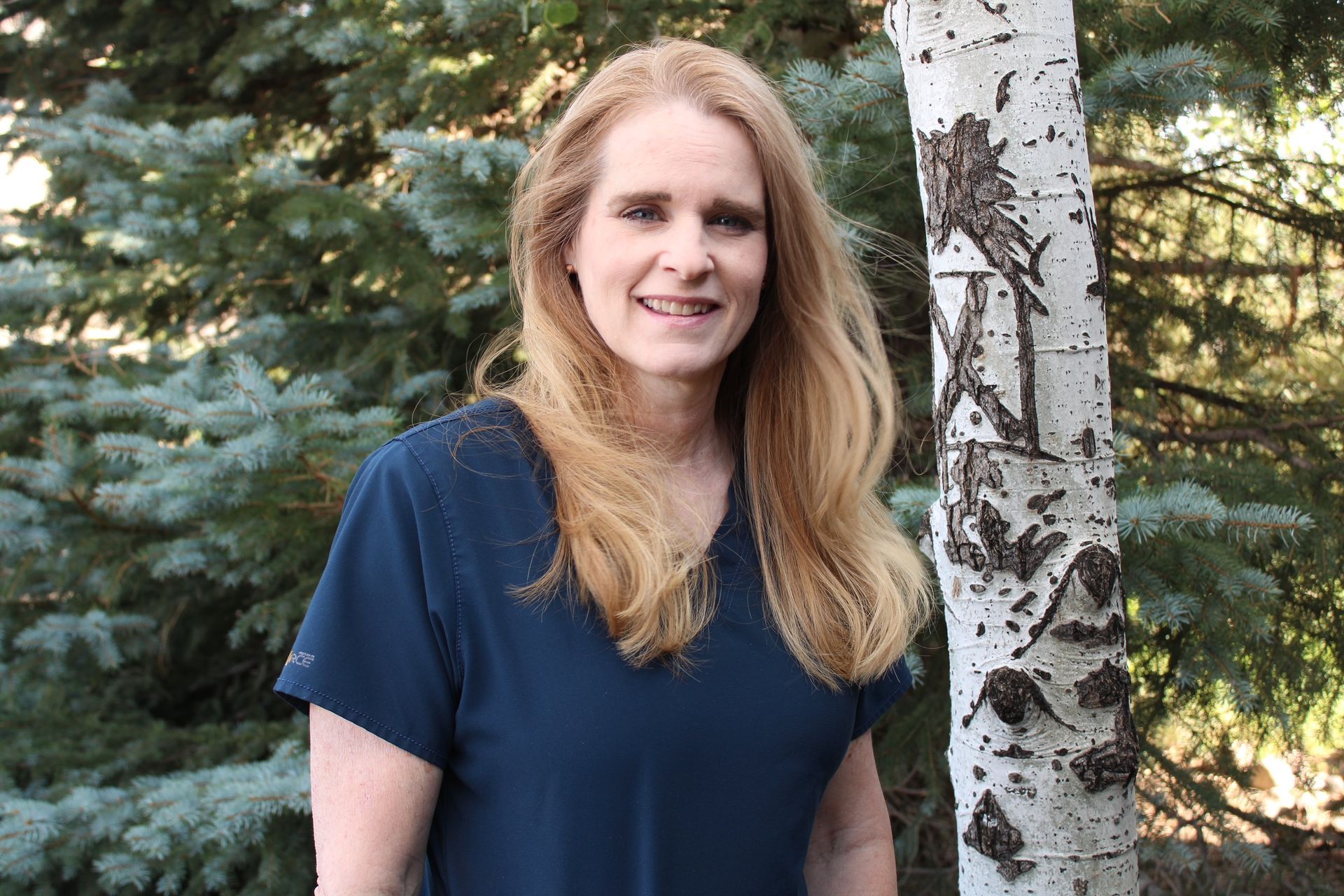 A woman in a blue scrub top is standing in front of a tree.