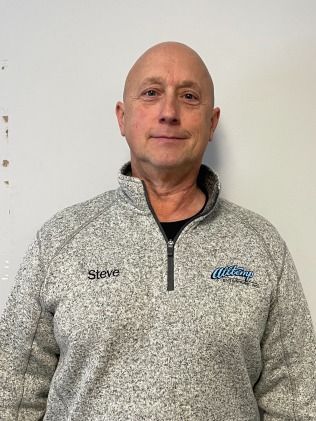Steve, a bald man, wearing a gray fleece with a zipper, smiling, standing in front of a white wall.