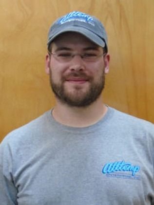 Man wearing a baseball cap and glasses, smiling. He has a beard and is wearing a gray t-shirt.