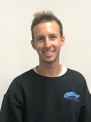 Young man with short brown hair, smiling, wearing a black sweatshirt with a logo, against a white wall.