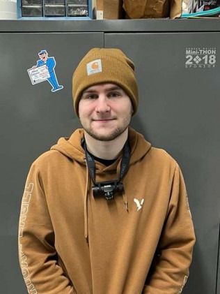 Young man in a brown beanie and hoodie smiles, wearing a headlamp, in front of a gray cabinet.