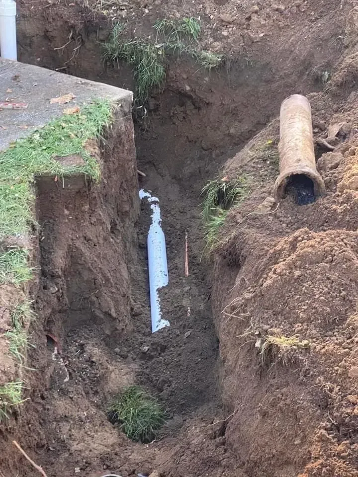 Trench dug in dirt, showing white PVC pipe and old corroded pipe.