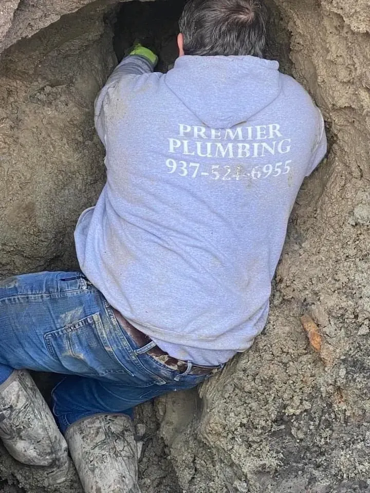 Plumber in light gray hoodie, jeans, and boots, working inside a hole in the ground.