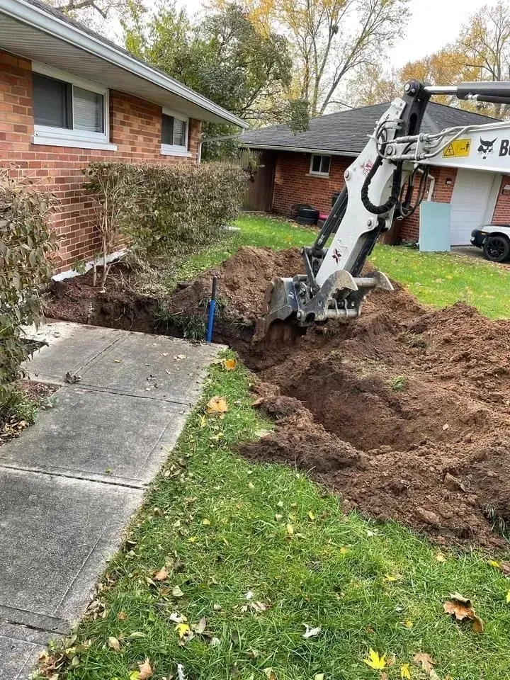 Excavator digging a trench next to a brick house and sidewalk.