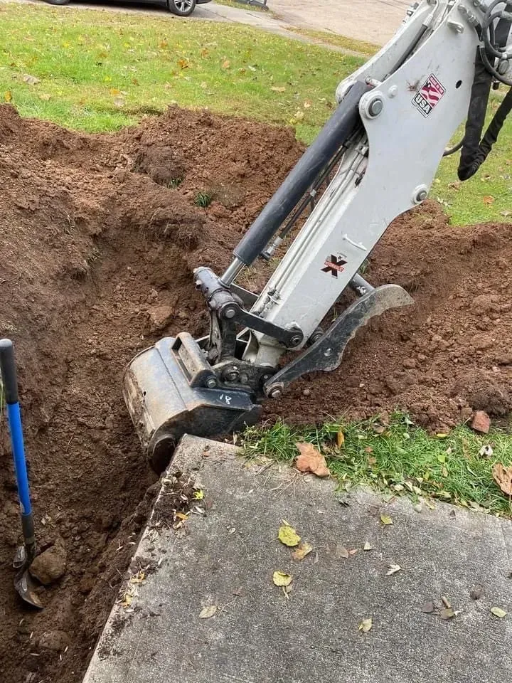Excavator digging a trench next to a concrete walkway. Dark soil, blue shovel nearby.