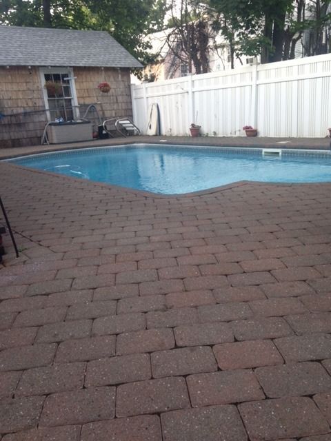 Backyard pool with brick patio, white fence, and small brick house in the background