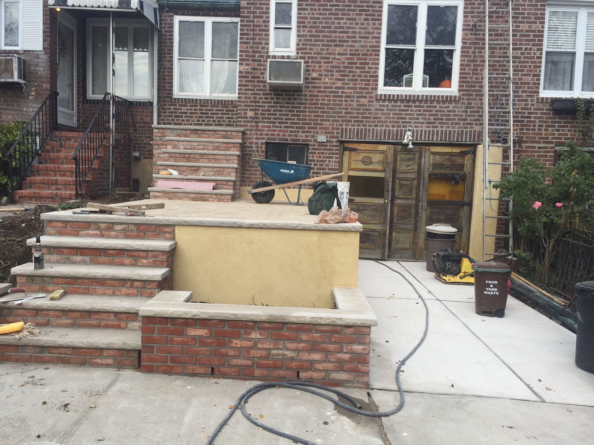 Backyard patio with brick steps, concrete walkway, chairs, and a small dog on the raised ledge.