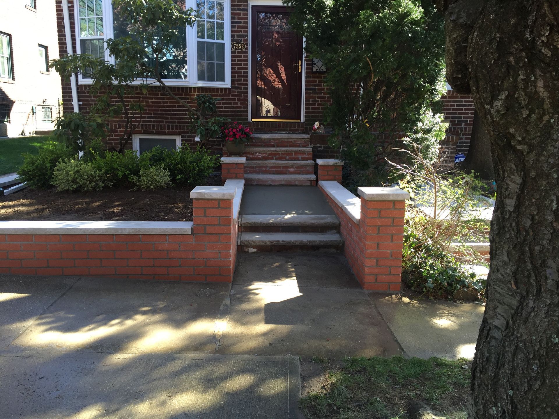 Brick house entrance with steps, short ramp, and brick side walls beside a tree-lined sidewalk.