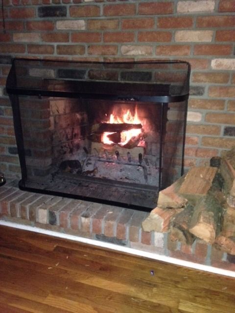 Lit fireplace with stacked firewood beside it in a brick-walled room.