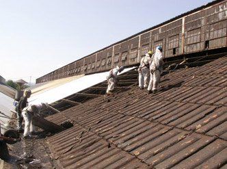 Workers in protective white suits remove corrugated roofing panels from an industrial building.
