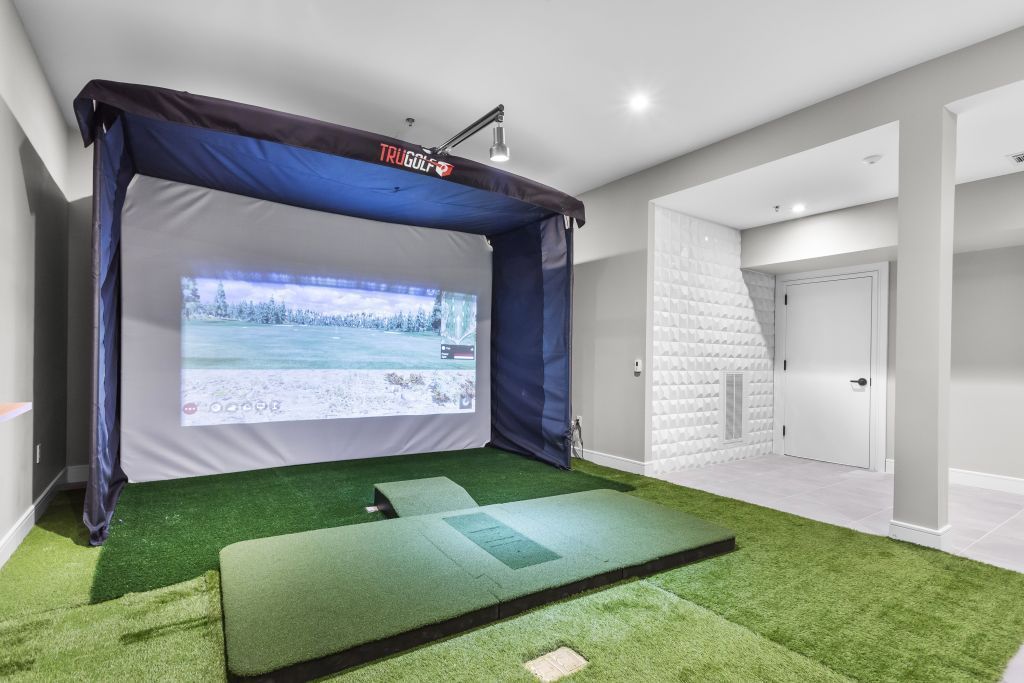 Indoor golf simulator room with green turf, large projection screen, and lounge area