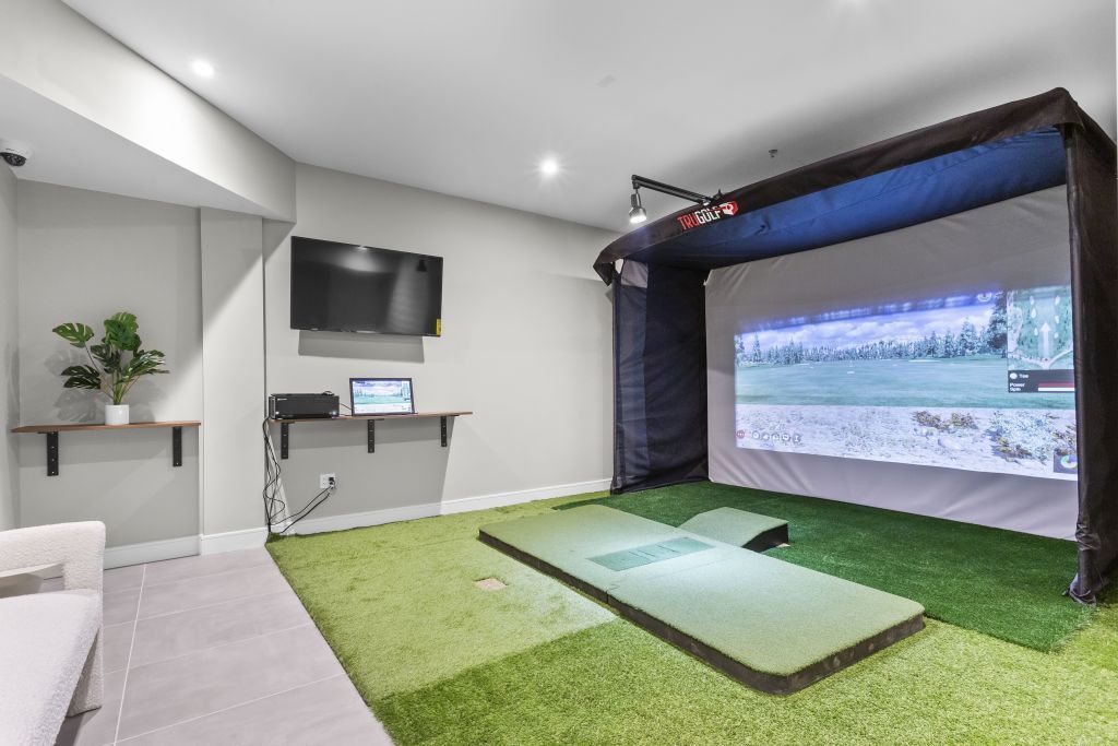 Indoor golf simulator room with turf mat, netted screen, wall-mounted TV, and lounge seating.
