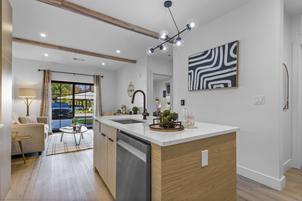 Bright modern kitchen with island, white countertops, wood floors, and living area beyond.