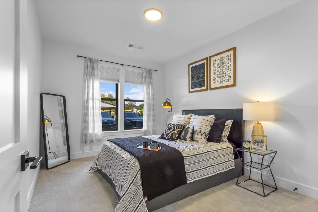 Bright modern bedroom with gray walls, patterned bedspread, lamp, mirror, and window view outdoors
