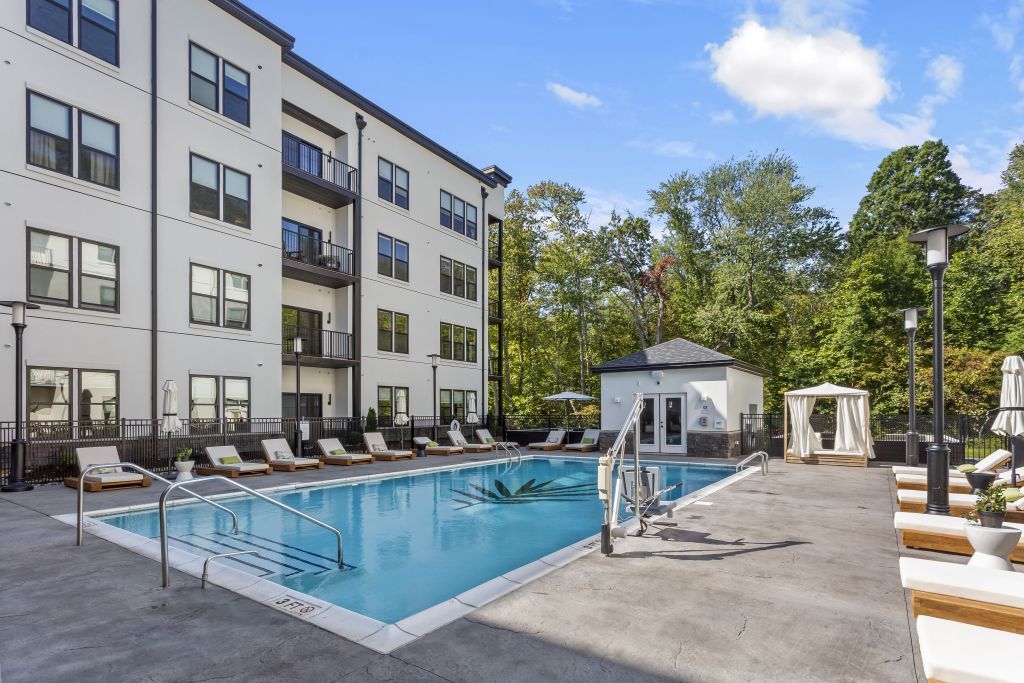 Modern apartment pool with lounge chairs, pergola seating, and trees on a sunny day