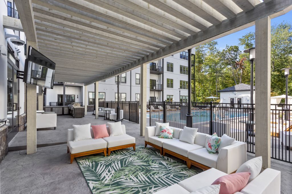 Modern outdoor lounge with white sofas, patterned rug, and a covered pergola beside apartment buildings