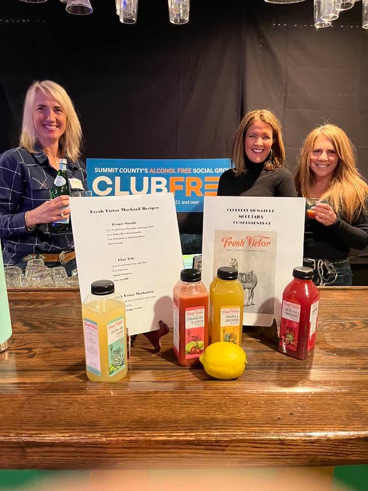 Three women smiling at a ClubFree event, standing behind a table laden with various containers of juice.