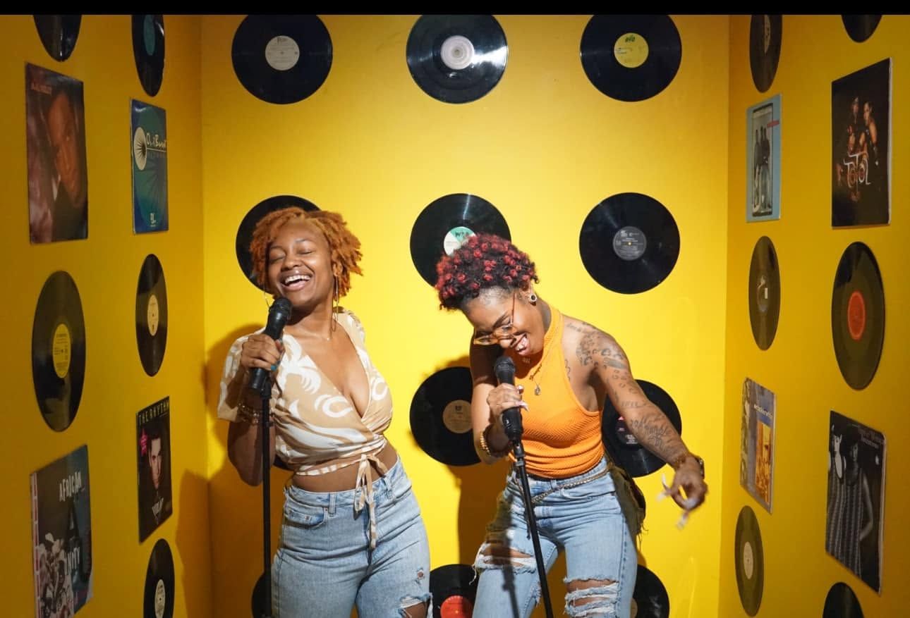 Two people singing into microphones in a yellow room with vinyl records on the walls.